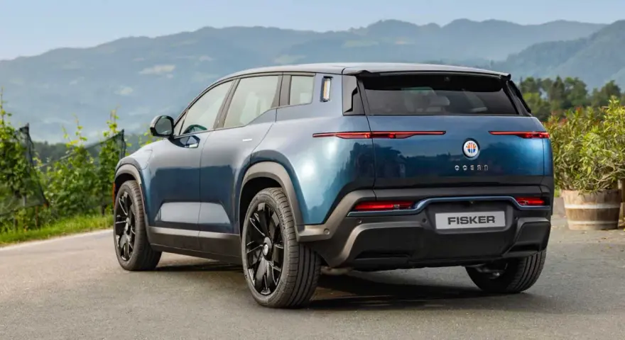 Tylna część niebieskiego elektrycznego SUV-a Fisker Ocean na tle gór i zieleni.