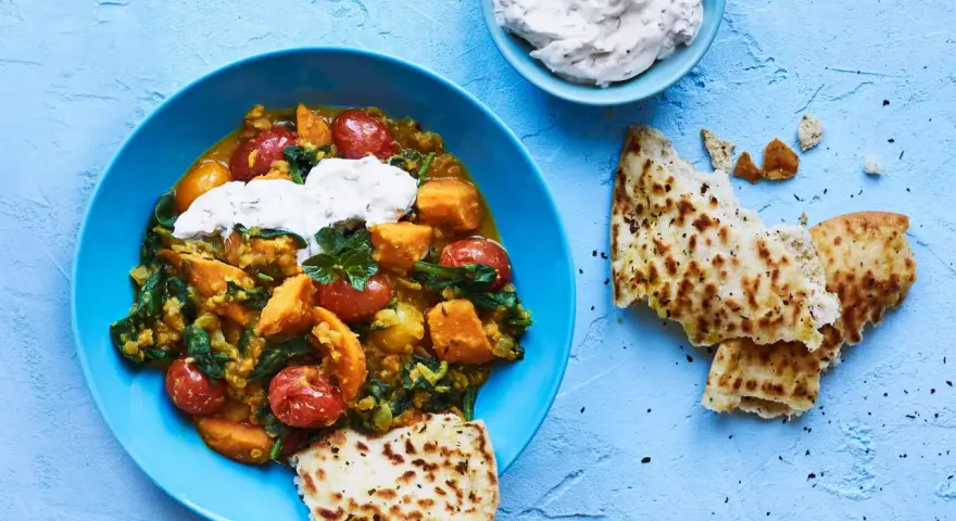 En blå skål med en mustig daal med sötpotatis, spenat och tomater, toppad med gräddfil. Serveras med naanbröd.