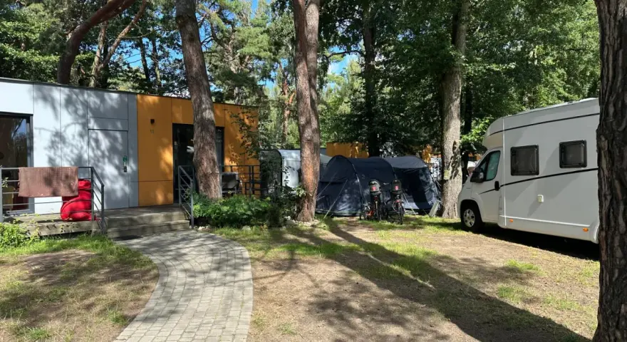 Na kempingu 45, wśród drzew, stoją nowoczesne domki, namioty i biała kamper.