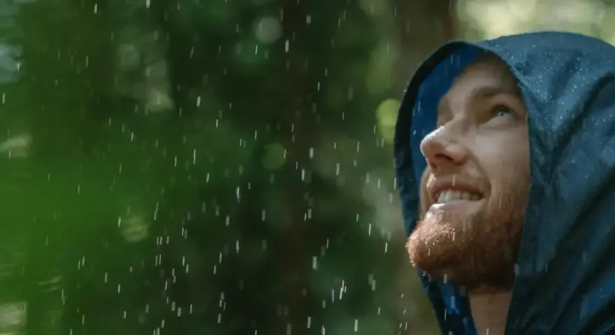 Mann mit Bart blickt lächelnd in den Regen, seine blaue Regenjacke hält ihn trocken.