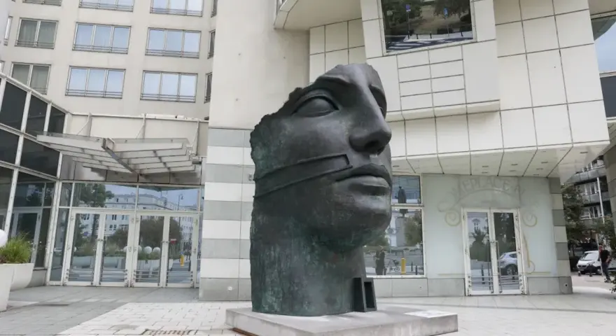 Okaleczona, ciemna rzeźba głowy z opaską na ustach, na tle nowoczesnego budynku.