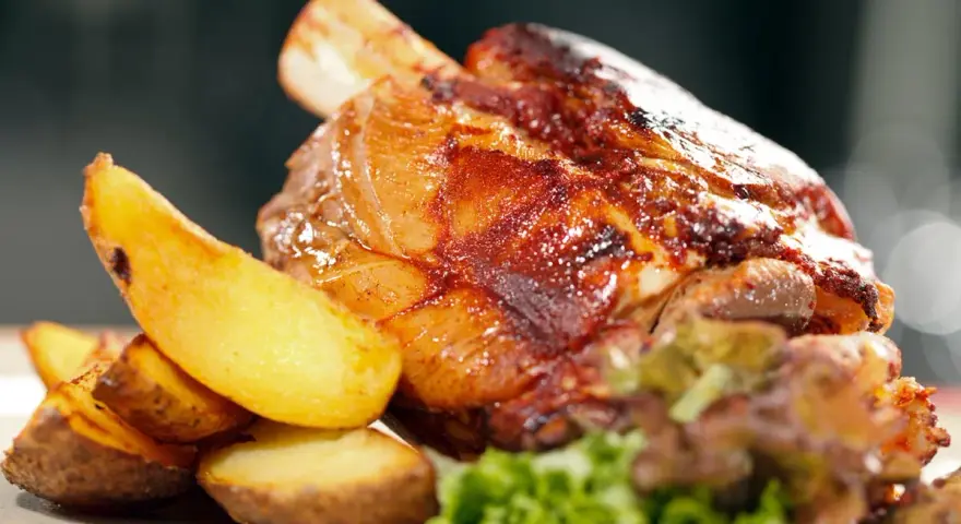 Codillo de cerdo asado, crujiente y jugoso, acompañado de patatas gajo doradas y una guarnición verde.