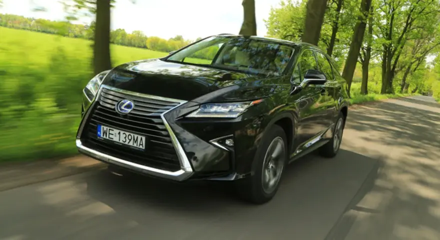 Czarny Lexus RX 450h pędzi po wiejskiej drodze, otoczony zielenią drzew i pól.