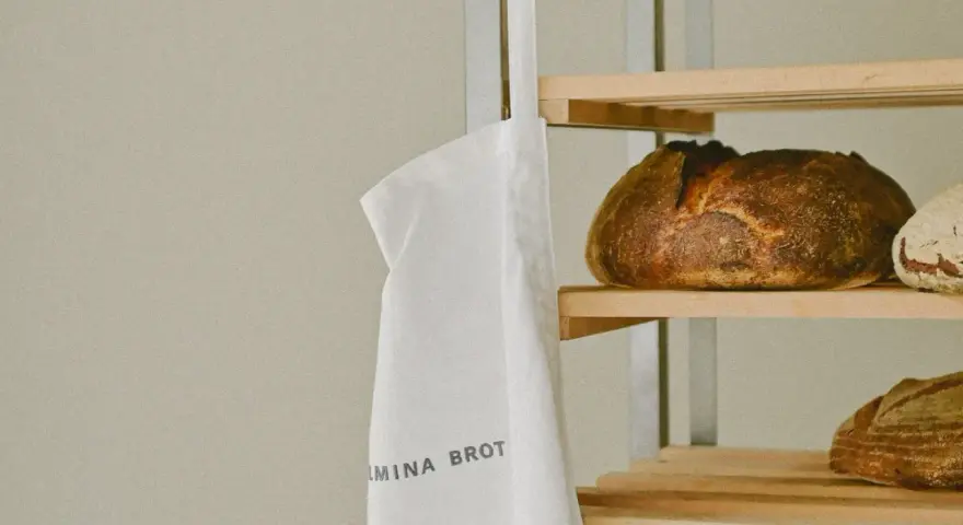 Ein weißer Stoffbeutel mit der Aufschrift "WILMINA BROT" hängt neben einem Regal mit frisch gebackenen Broten.