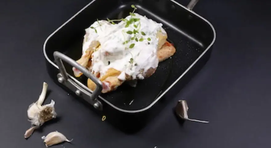 Un poulet entier est nappé de fromage blanc crémeux et garni d'herbes fraîches dans un plat noir. Des gousses d'ail sont disposées autour.