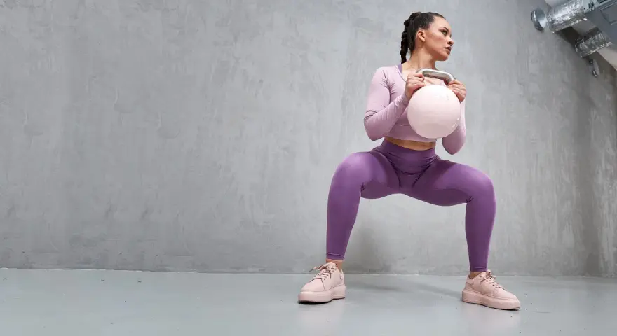 En kvinna i lila träningskläder gör en kettlebell swing-rörelse, med en ljusrosa kettlebell i händerna.