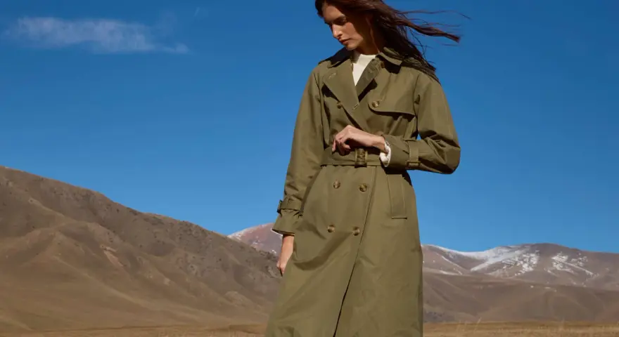 Mujer con un elegante trench color caqui, con el pelo al viento, en un paisaje montañoso bajo un cielo azul.