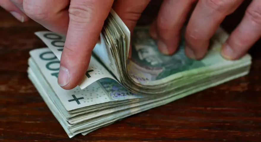 Dłonie przewracają stos banknotów 100 zł na drewnianym blacie.
