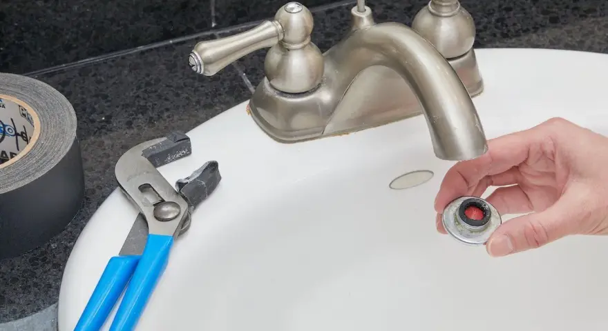 Hand holding a removed faucet aerator with red buildup, next to pliers and duct tape on a sink.