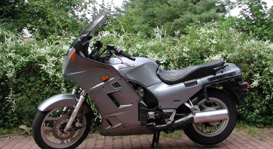 Srebrny motocykl Kawasaki GTR 1000 zaparkowany na czerwonej kostce brukowej, otoczony zielenią.