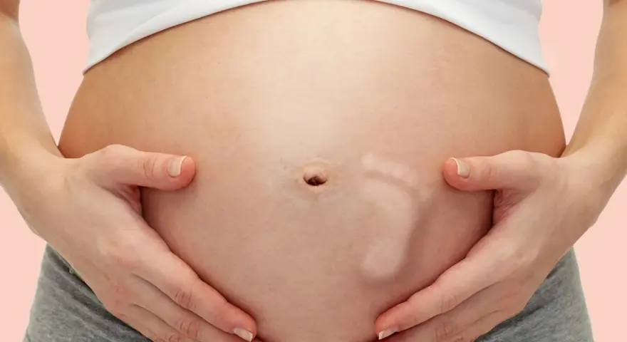 Brzuszek w 30 tygodniu ciąży, z widocznym odciskiem stópki dziecka. Ręce mamy delikatnie obejmują rosnący brzuch.