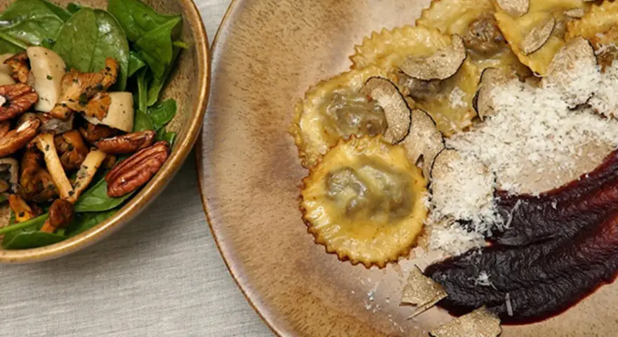 Ravioli pasta mit Trüffeln, Parmesan und einer dunklen Sauce. Daneben eine Schüssel mit Pilzen, Nüssen und Spinat.