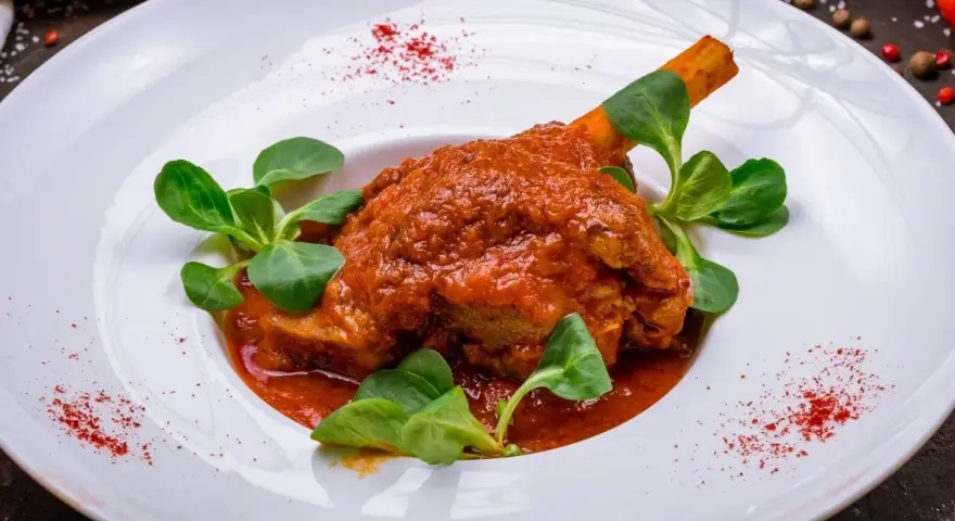 Pierna de cordero en salsa roja con hojas verdes en plato blanco.