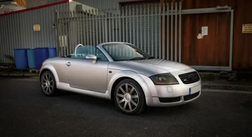 Silberner Audi TT 2002 Roadster mit offenem Verdeck steht auf einer Straße vor einem Industriegebäude.
