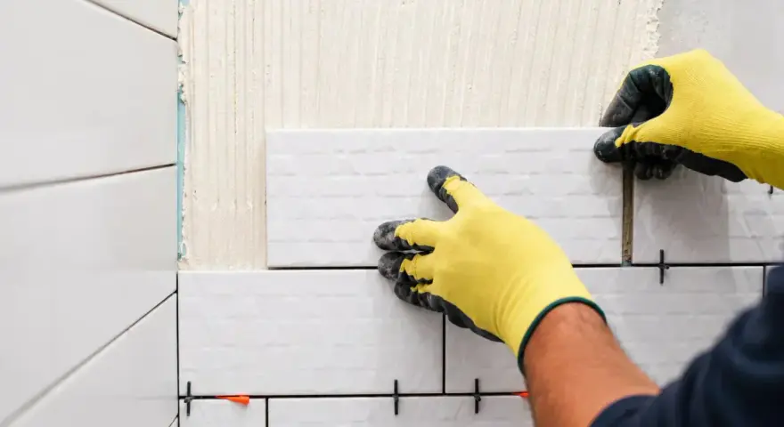 Mains gantées de jaune en train de coller du carrelage sur du carrelage, avec des croisillons pour un espacement parfait.