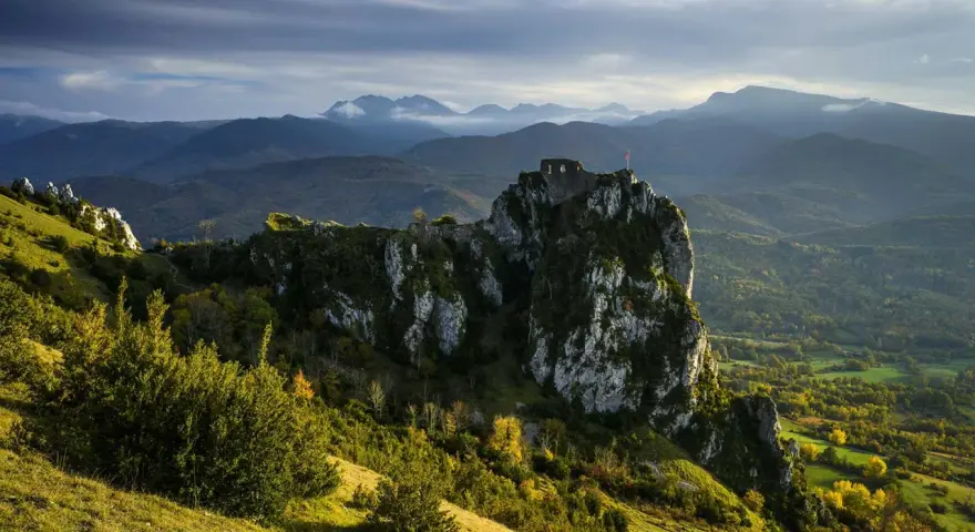 Ruines d'un château cathare sur un pic rocheux, dominant un paysage de montagnes et de forêts. Le chemin des bonshommes serpente à travers la vallée.