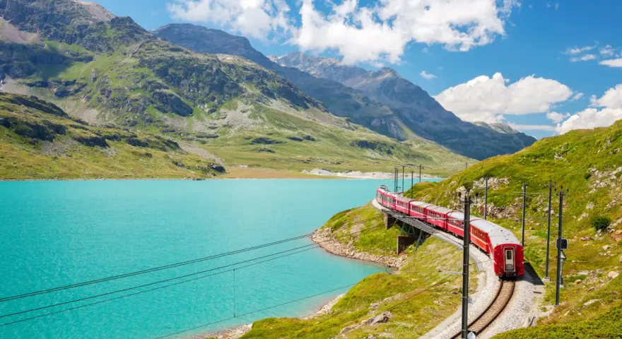 A red train winds along a track beside a vibrant turquoise lake, with lush green mountains and a bright blue sky in the background. This is a classic como to switzerland train journey.