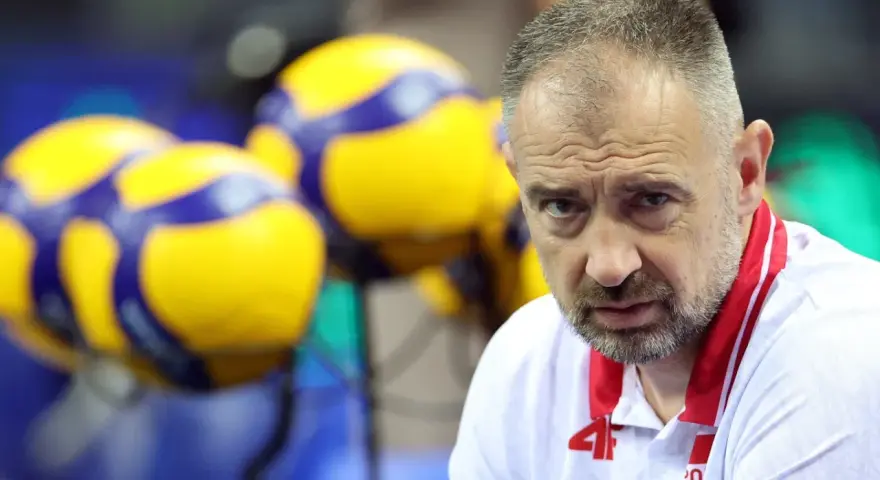 Skupiony Nikola Grbić, trener siatkówki, w białej koszulce z czerwonymi akcentami, na tle żółto-niebieskich piłek.