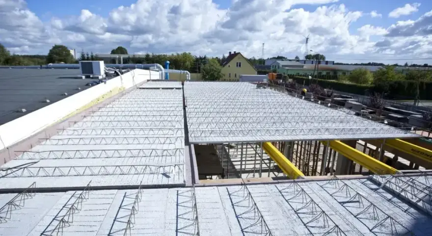 Budowa dachu kopertowego z elementami kratownicowymi i betonem. W tle budynki i błękitne niebo z chmurami.