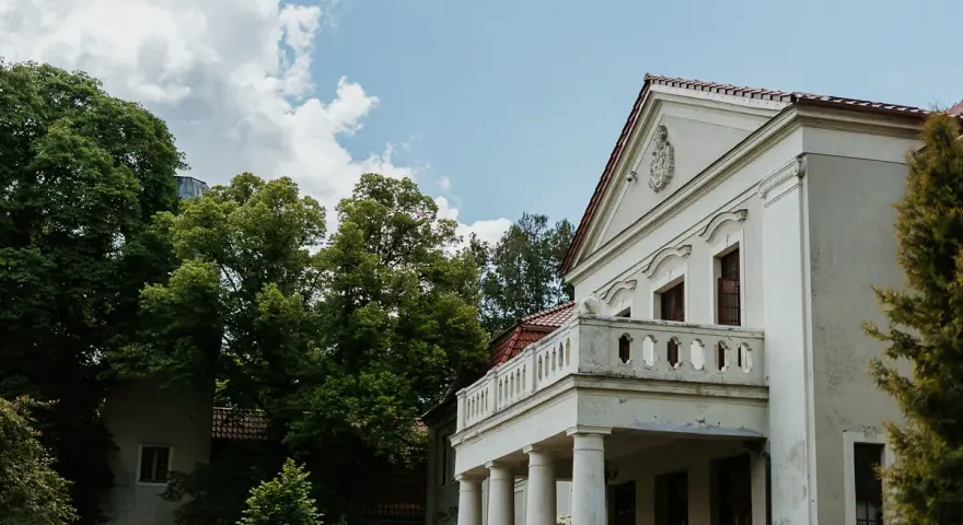 Pałac Grąbkowo, biały budynek z kolumnami i balkonem, otoczony bujną zielenią drzew pod błękitnym niebem z chmurami.