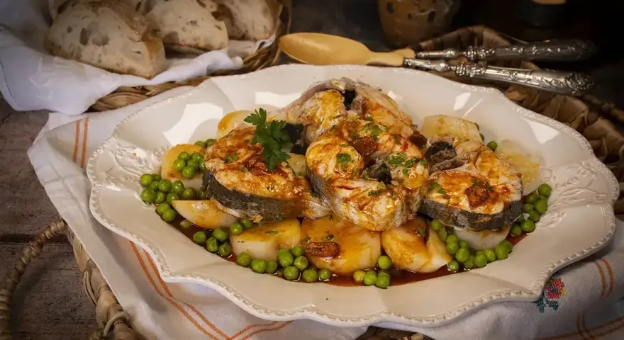 Merluza a la gallega servida en plato blanco con patatas, guisantes y perejil. Pan rústico en cesta al fondo.