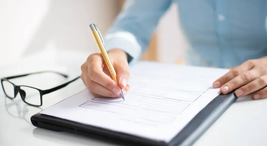 Ręka pisze złote pióro na dokumencie, który może być wzorem wniosku o opiekę nad dzieckiem. Obok leżą okulary.