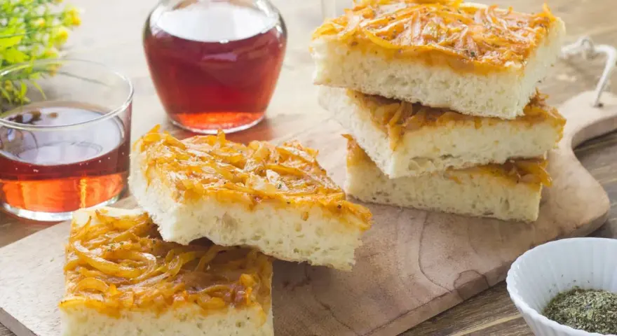 Focaccia con cipolle dorate e soffice, servita su un tagliere di legno con un bicchiere di vino rosato.