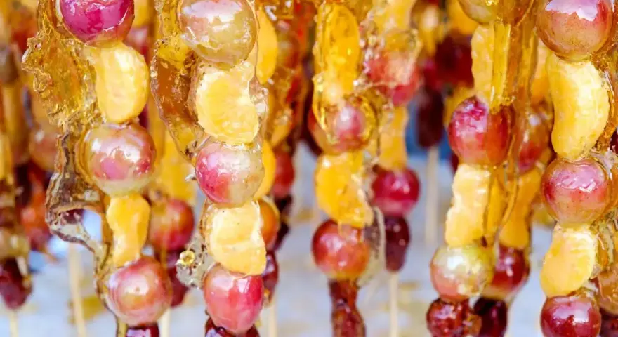 Spiedini di frutta caramellata con acqua e zucchero, con uva e spicchi d'arancia lucidi e dorati.