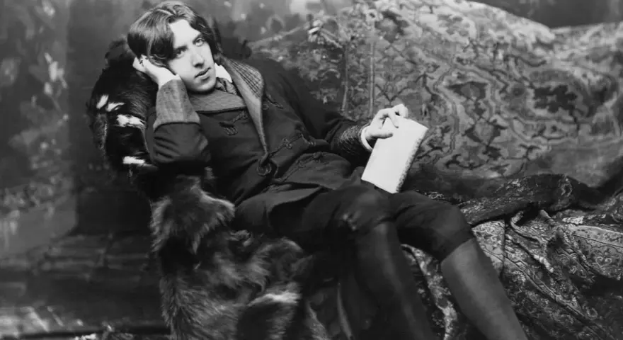 Oscar Wilde, w futrzanym płaszczu, z książką w dłoni, w pozie sugerującej refleksję nad swoimi sztukami.