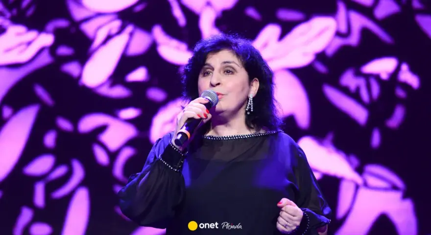 Kobieta śpiewa na scenie, w czarnej, ozdobnej sukni. Wiek męża Eleni nie jest widoczny na zdjęciu.