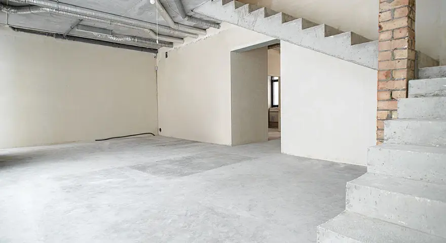 Wnętrze w stanie deweloperskim: betonowe schody, nieotynkowane ściany, widoczne instalacje i przewody.
