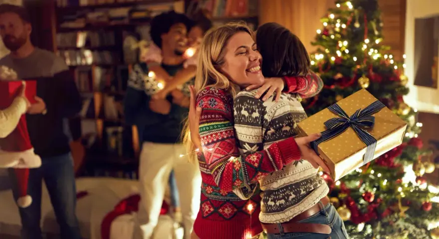 Zwei Frauen umarmen sich vor einem Weihnachtsbaum, eine hält ein Geschenk. Eine Familie feiert Weihnachten, eine der vielen schönen weihnachtstraditionen deutschland.