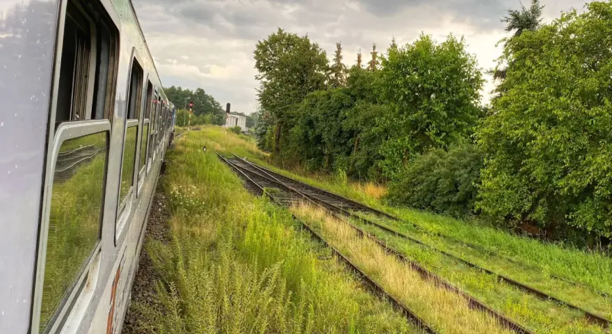 Widok z okna pociągu na tory kolejowe otoczone zielenią i chmury na niebie.