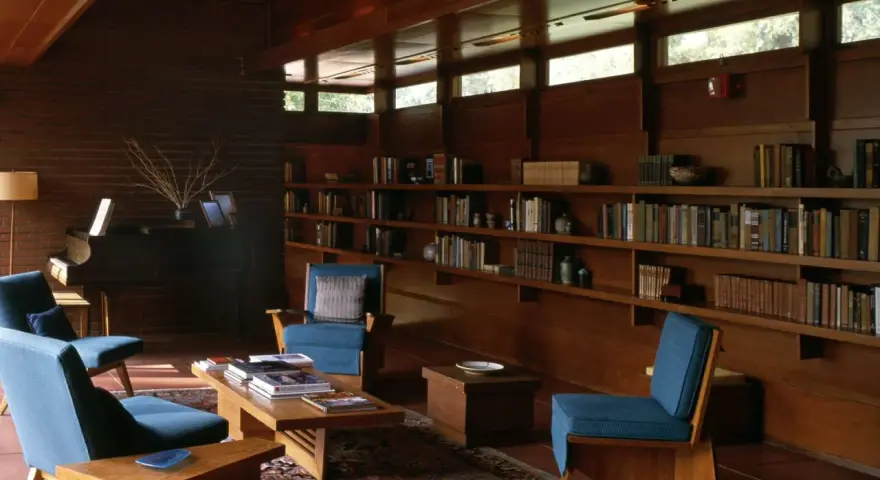 A cozy living room with blue chairs, a piano, and bookshelves. The clerestory windows above let in natural light, illuminating the warm wood interior.