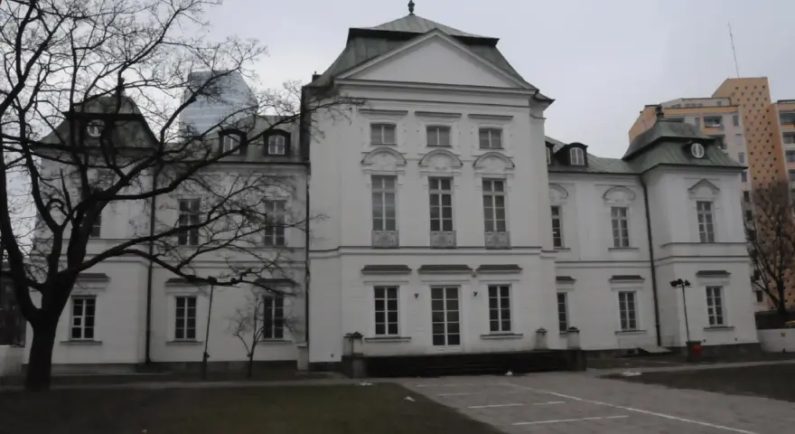 Pałac Przebendowskich, biała, elegancka budowla z zielonym dachem, otoczona drzewami i nowoczesnymi budynkami.
