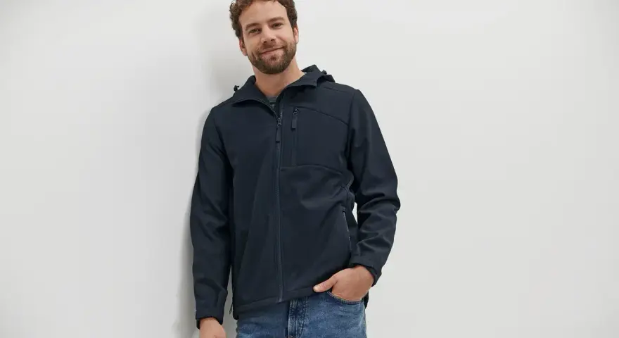 Mann mit lockigem Haar trägt eine dunkelblaue Softshelljacke und Jeans. Perfekt für das nächste Abenteuer oder zum Softshelljacke waschen.