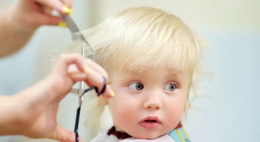 Ein kleines blondes Kind bekommt die Haare geschnitten. Die Schere nähert sich vorsichtig den feinen Strähnen.