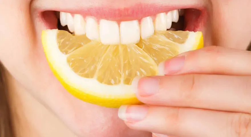 Une personne tient une tranche de citron près de sa bouche ouverte, révélant des dents blanches. Le citron est connu pour ses bienfaits sur la peau.