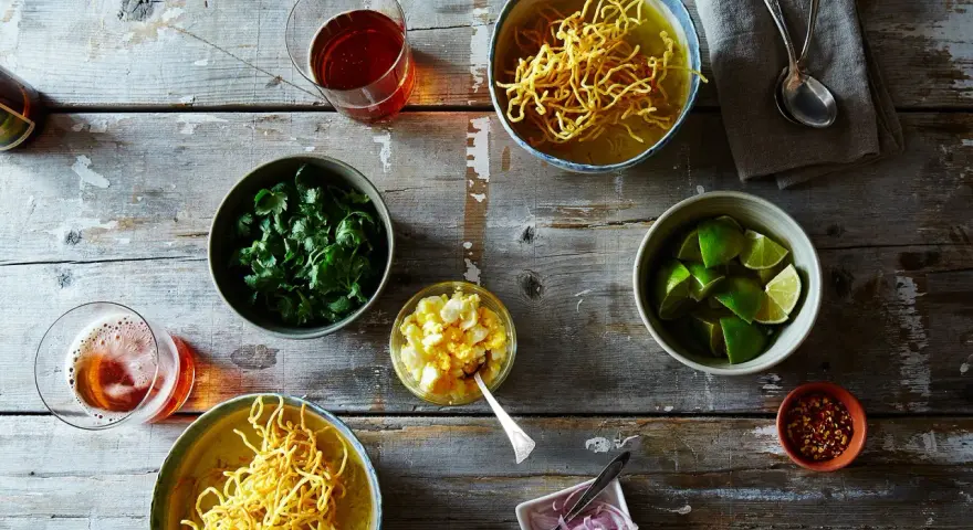 Dwa miseczki khao soi, świeża kolendra, limonki, jajko na twardo, czerwona cebula i płatki chili na drewnianym stole.