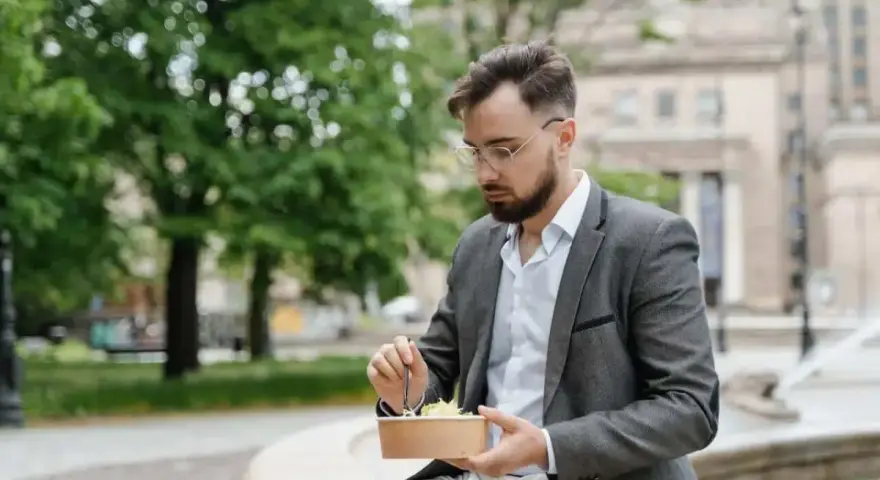 Mężczyzna w okularach i marynarce robi sobie przerwę w pracy, jedząc sałatkę na ławce w parku.