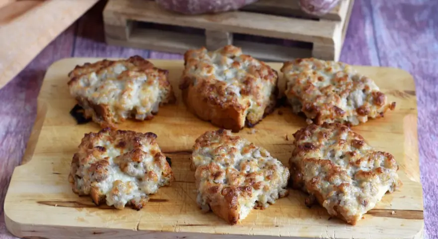 Sei crostini salsiccia e stracchino dorati e filanti, pronti per essere gustati su un tagliere di legno.