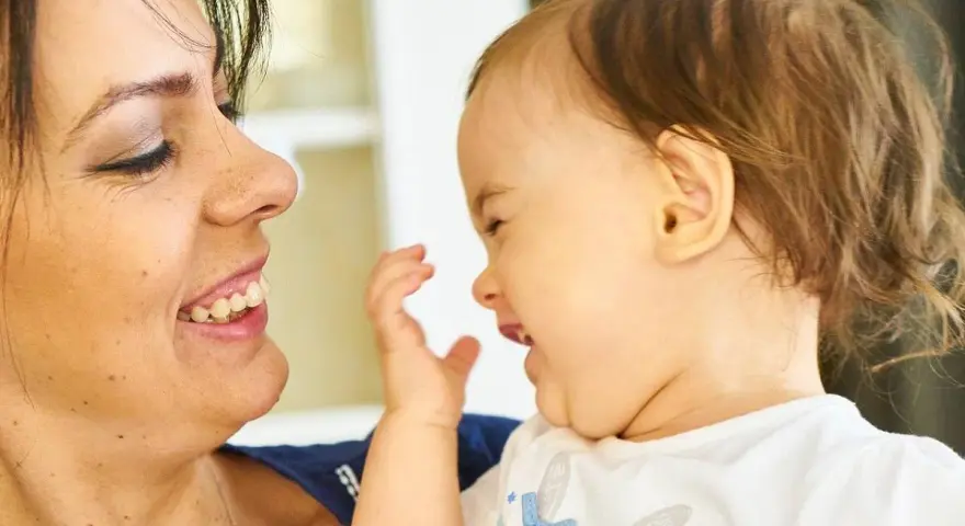 Uśmiechnięta mama patrzy na swoje dziecko, które bawi się i śmieje, wyciągając rączkę.
