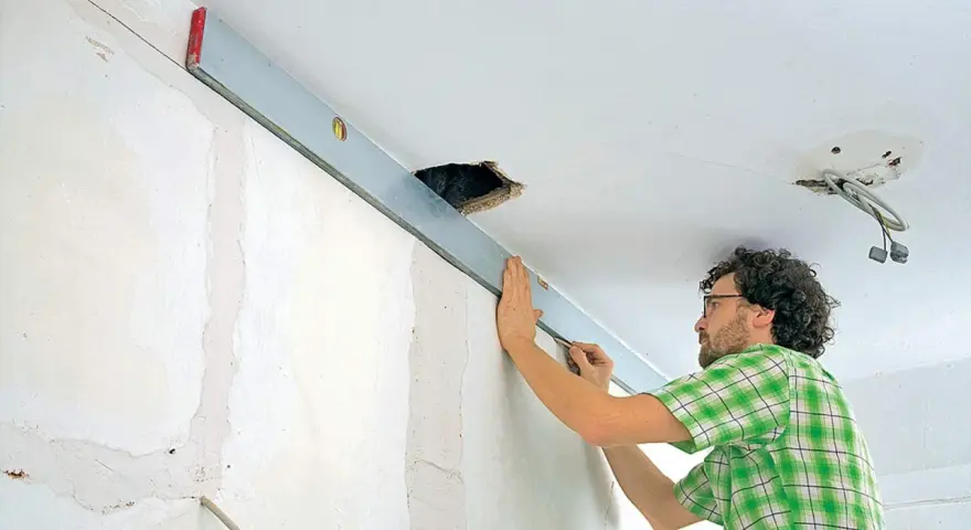 Mann richtet eine Wasserwaage an der Decke aus, um die Wand mit Rigips zu verkleiden. Kabel hängen aus einer Deckenöffnung.
