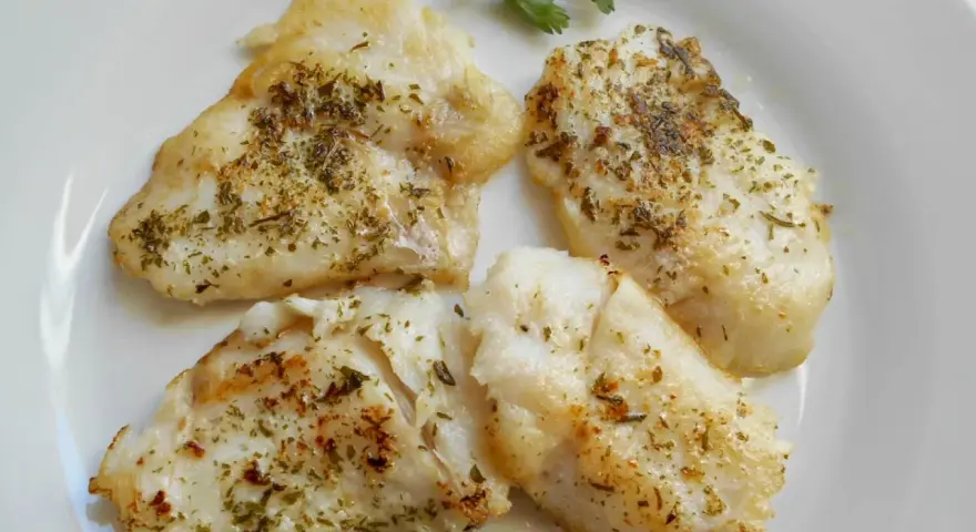 Cuatro trozos de pescado blanco rosada a la plancha, sazonados con hierbas, servidos en un plato blanco con una ramita de perejil.