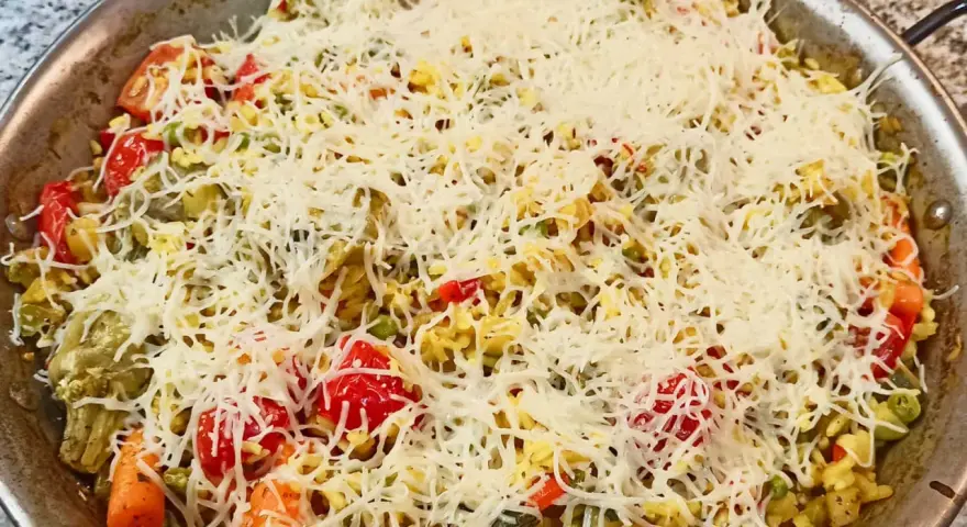 Arroz a la milanesa listo para hornear, cubierto con queso rallado y vegetales frescos como tomates, zanahorias y guisantes.