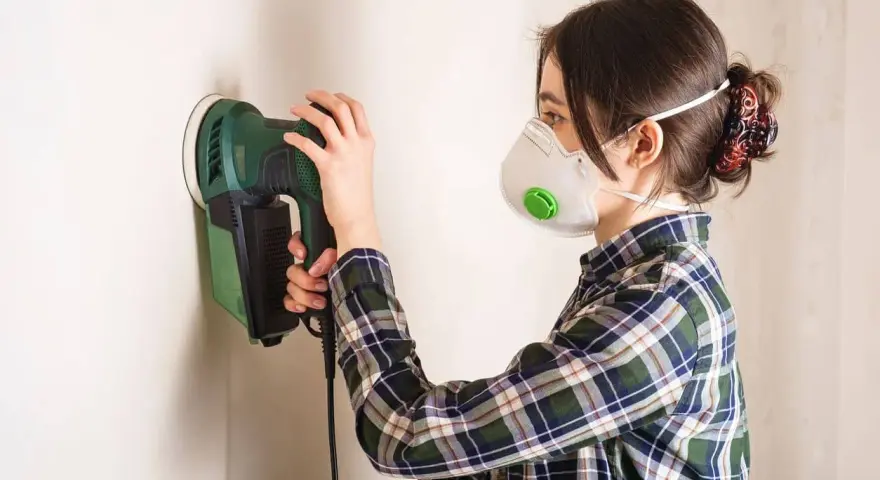 Persona con mascarilla y camisa a cuadros usa una lijadora eléctrica para lijar una pared.