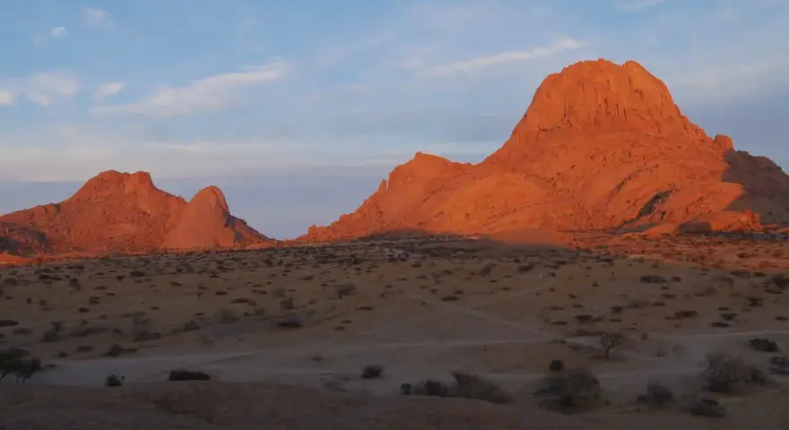 Zachód słońca maluje na pomarańczowo skalne szczyty Spitzkoppe w Namibii. Pustynny krajobraz z suchymi krzewami i ścieżkami.