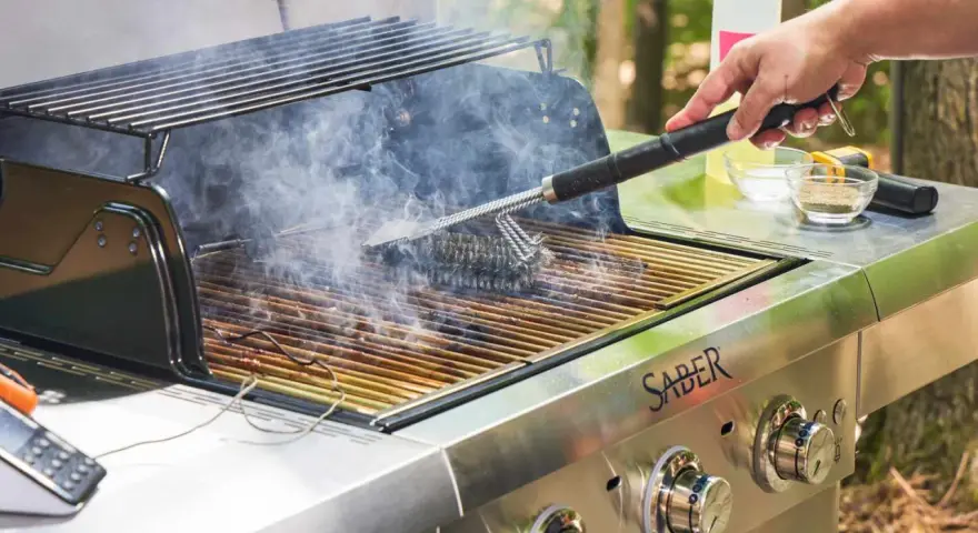 A hand uses a grill brush to clean the grates of a smoking Saber grill, showing the best way to clean a grill for your next cookout.