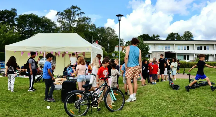 Barn och vuxna samlas vid ett tält på en gräsmatta, kanske för att dela ett hemligt språk. En cykel och elsparkcyklar syns.