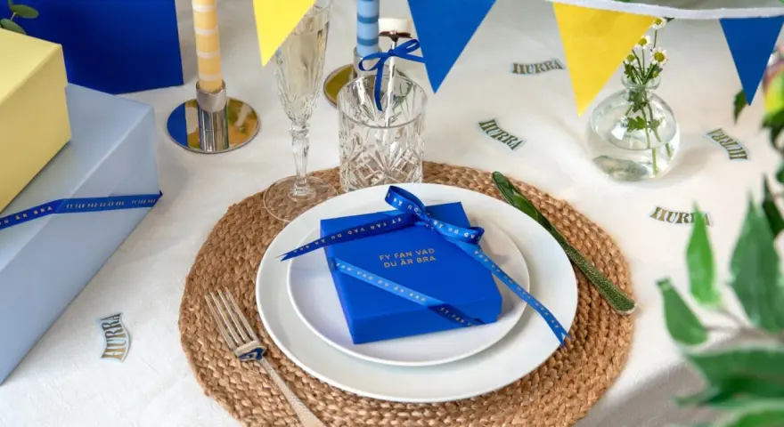 Blå och gula girlanger, ljus och presenter på ett dukat bord. En blå present med band och texten "Fy fan vad du är bra" är en fin pom pom dekoration.